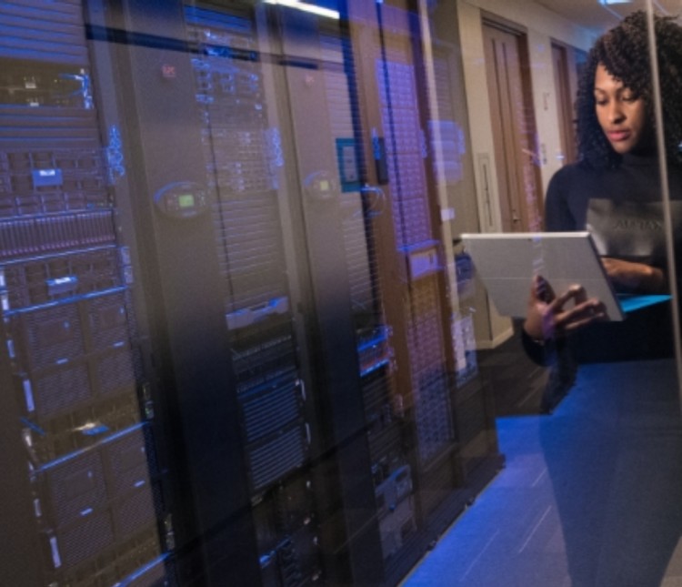Image of a woman holding a laptop while standing beside a server room representing the ease of migrating device automatically through IT automation.
